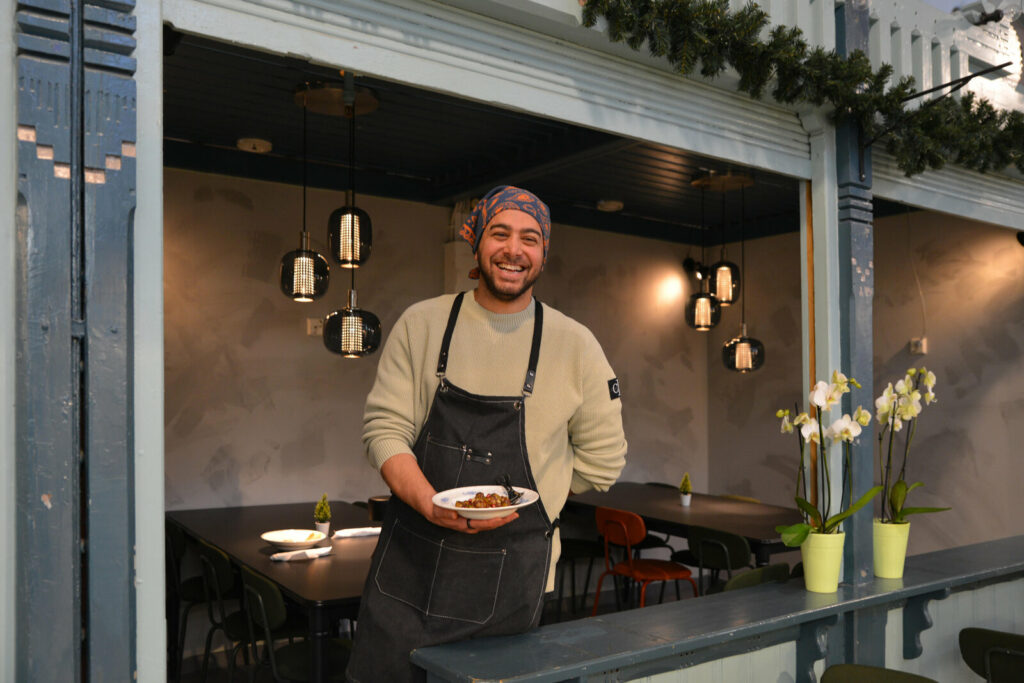 Maahan Delin yrittäjä Maahoo seisoo ruokailutilan edessä annoslautanen kädessään ja hymyilee leveästi.